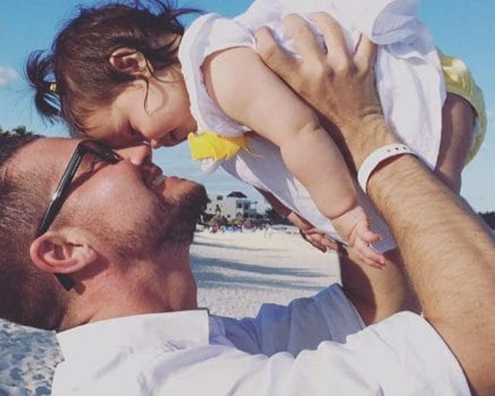 A man holding his baby daughter in the air making their foreheads touch at a beach.