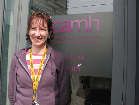 A woman with ginger hair and bangs wearing a half zipped purple hoodie, smiling with her hands clasped behind her back. 