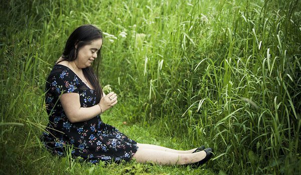 A woman with down syndrome wearing a black floral dress, sitting in a green field looking at a flower in her hand. 