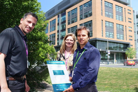 Two CAMH staff standing in a field in front of the Bell Gateway Building with a man in a green lanyard holding a blue and white plaque. 