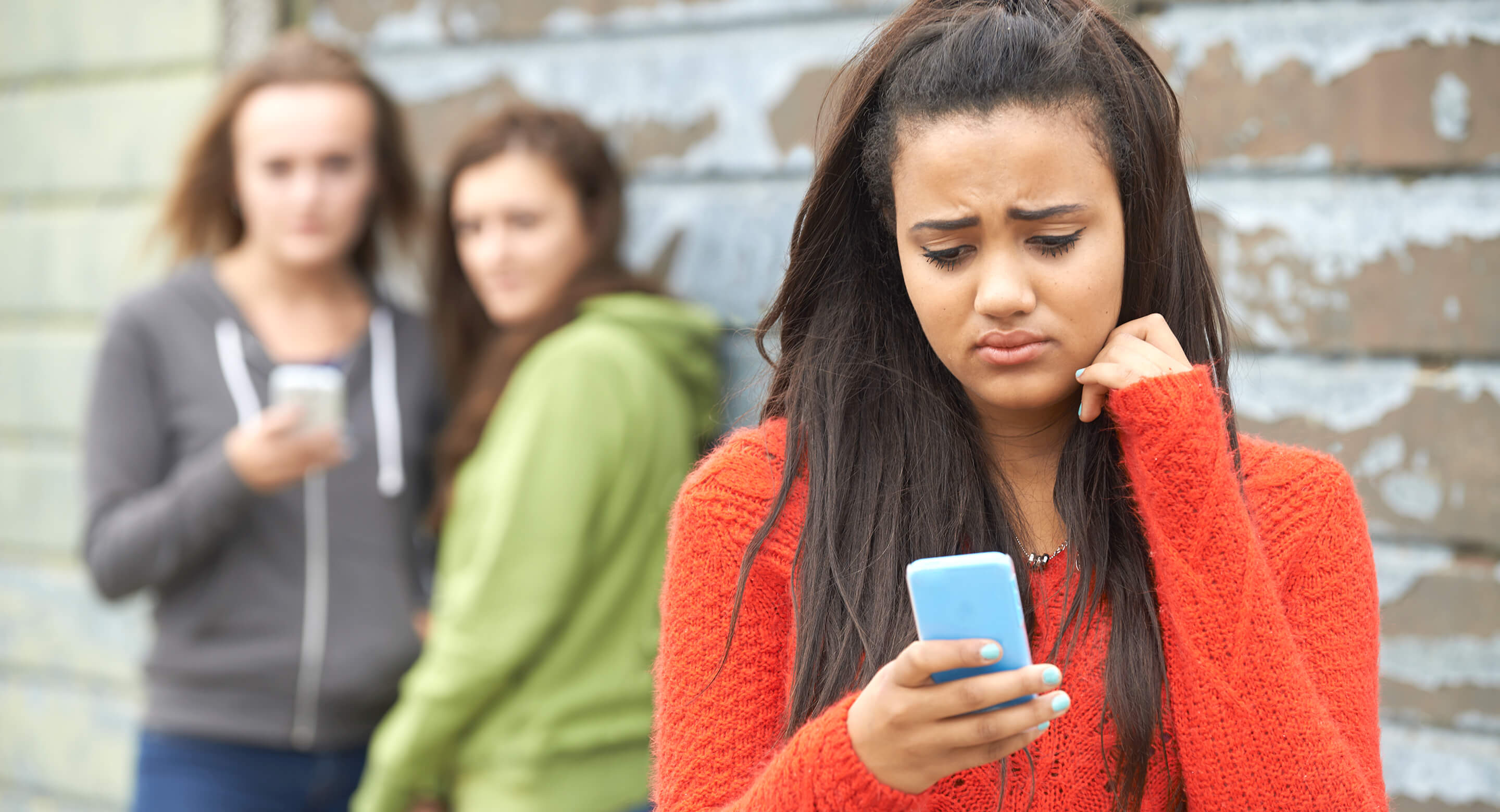 Girls looks sadly at her phone with other girls gossiping in background