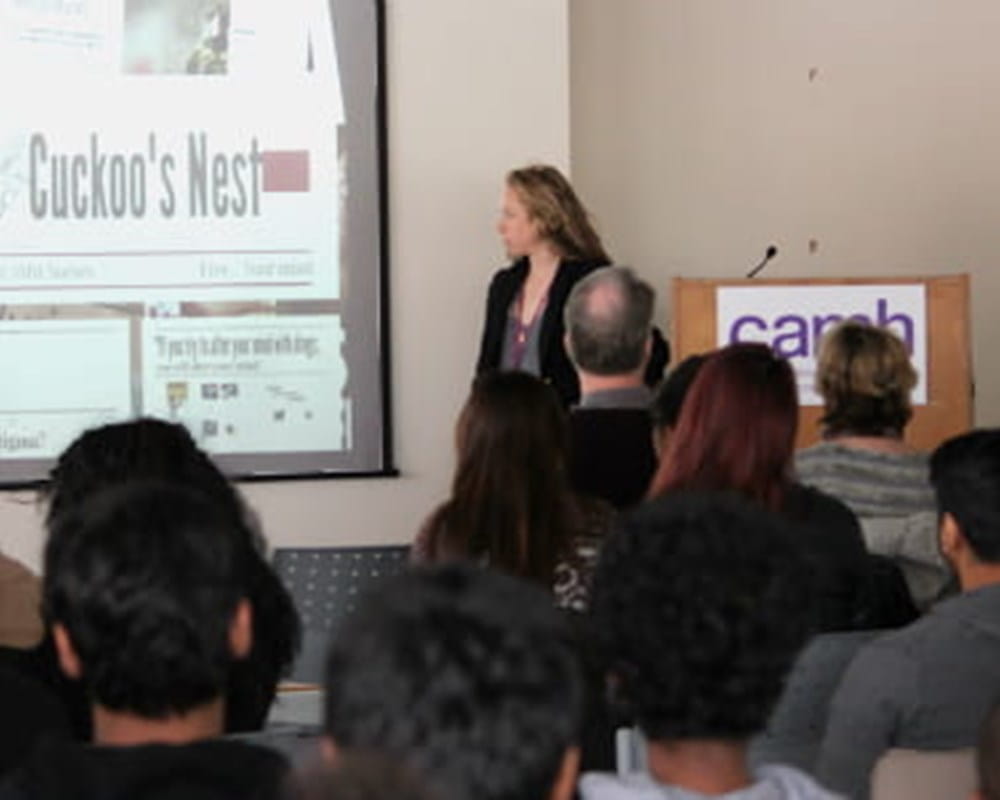 A woman presenting in front of a large diverse crowd with a projected image of a newspaper headline
