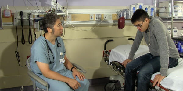 A patient sitting on a hospital bed and a doctor in green scrubs talking to him. 