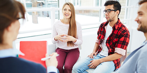 A woman with long hair and two men with facial hair sitting around a woman with classes and a red notebook. 