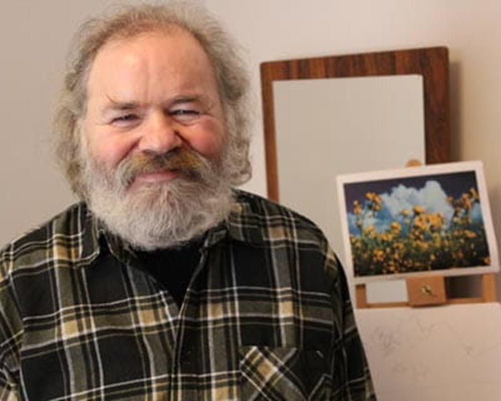 An older man with grey hair and a grey beard in a plaid shirt smiling at the camera.