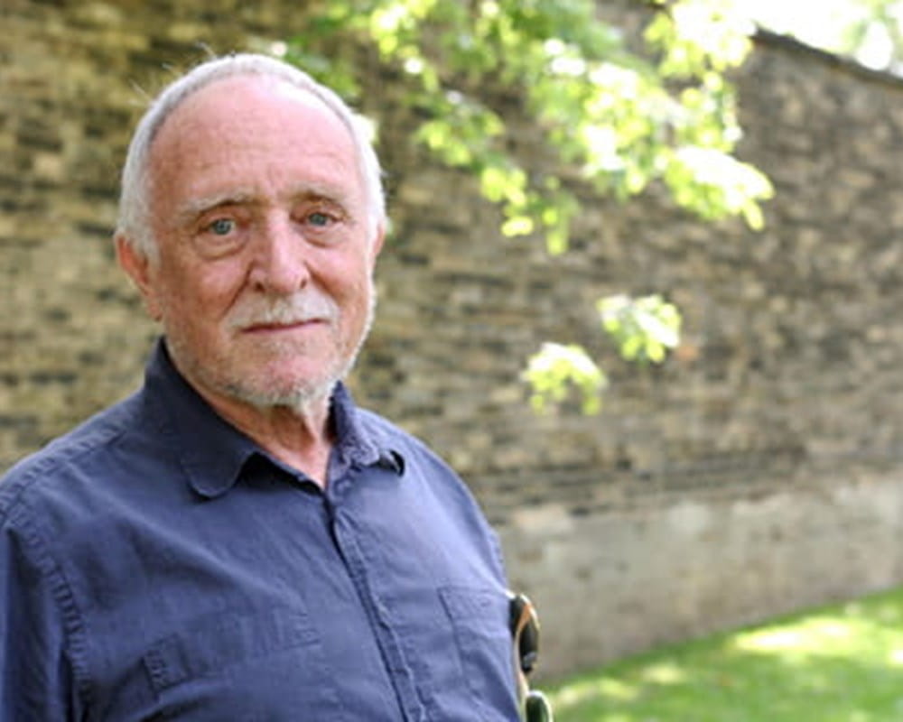 An older man with white hair wearing a blue button up standing in front of a brick wall, smiling for the camera.