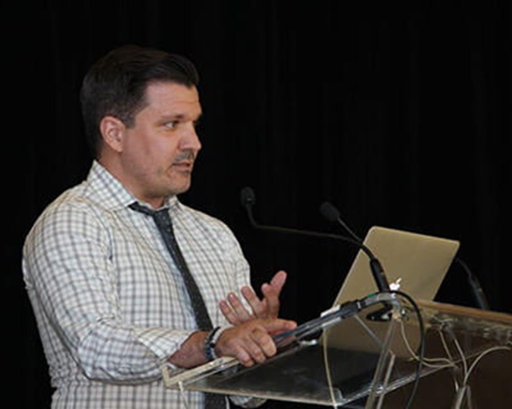 A man in a white and grey checkered button shirt and black tie speaking in front of a clear podium.