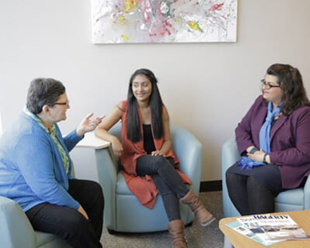 Three female students sitting on blue chairs and having a conversation.